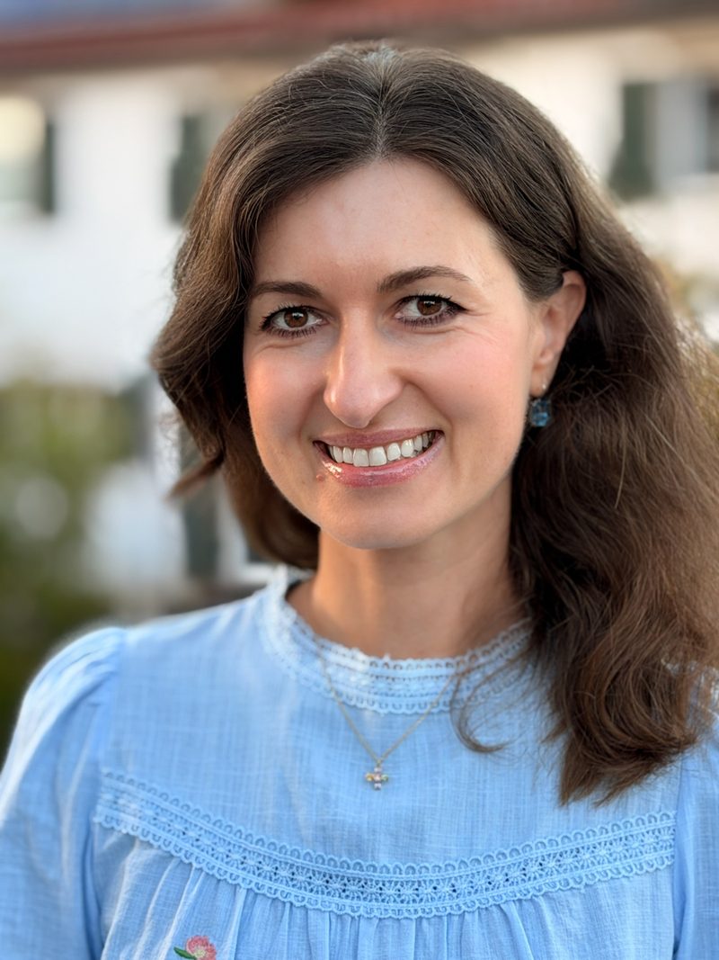Diana Vornicu-Eger, lächelnd, mit langen welligen Haaren, in einer hellblauen Bluse mit Spitze, draußen im warmen Abendlicht.