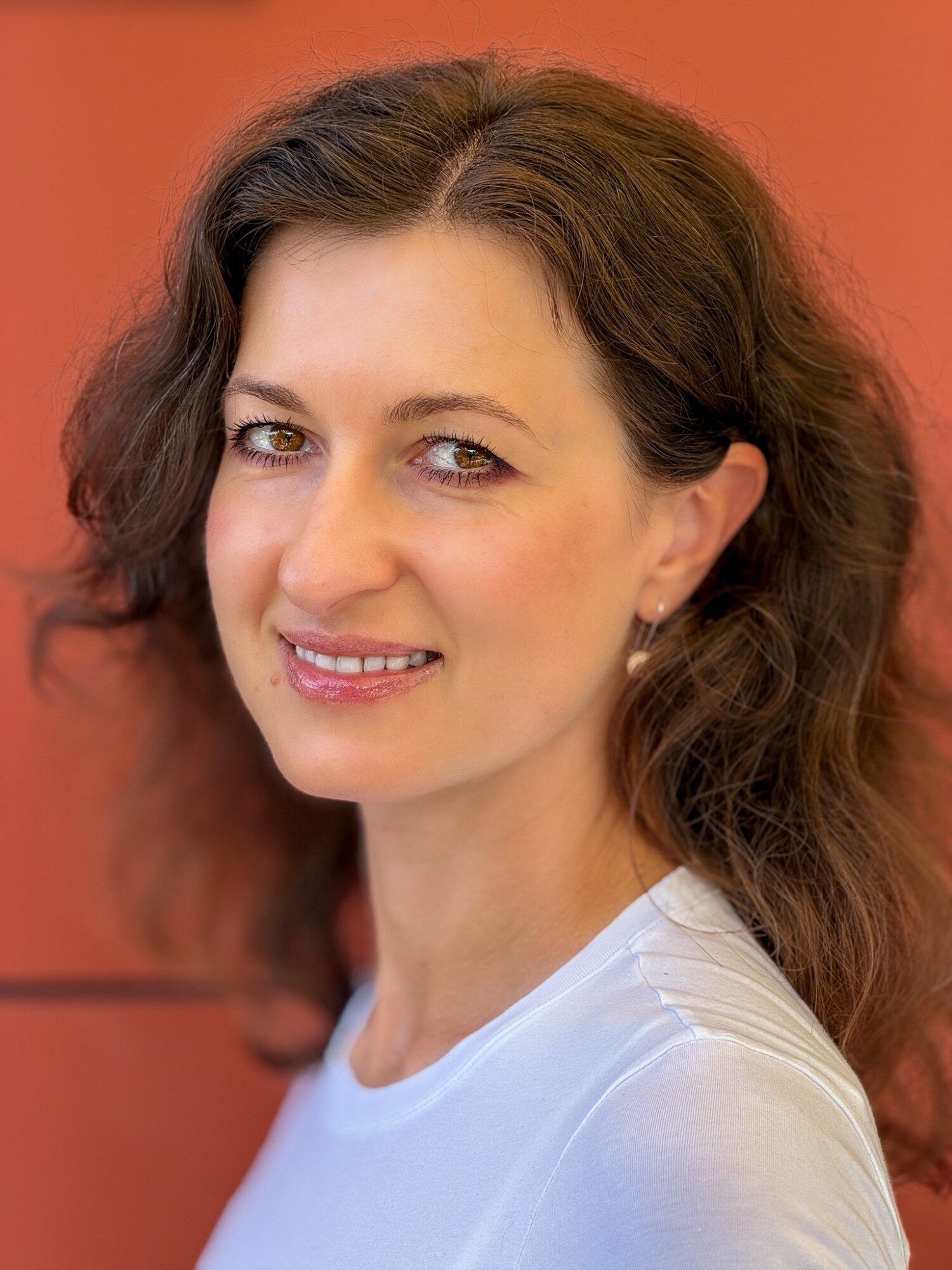 Diana Vornicu-Eger, lächelnd, mit langen welligen Haaren, in einem weißen T-Shirt vor einer terrakottafarbenen Wand.