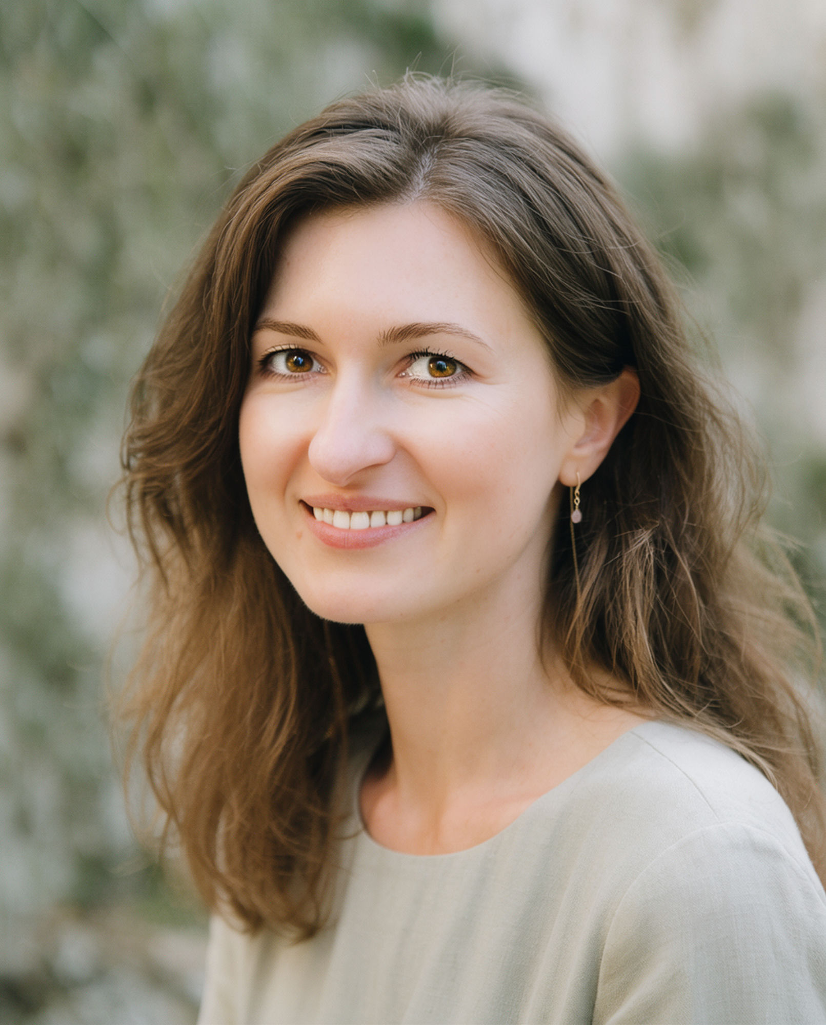 Diana Vornicu-Eger, lächelnd, mit langen welligen Haaren, in einer salbeifarbenen Bluse vor weichem grünem Hintergrund.
